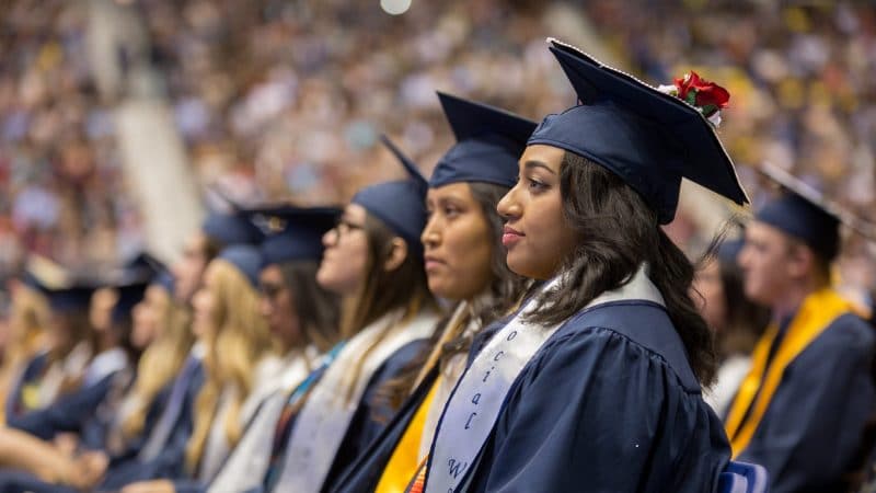 Students in graduation gowns and caps sit in a row in auditorium.
