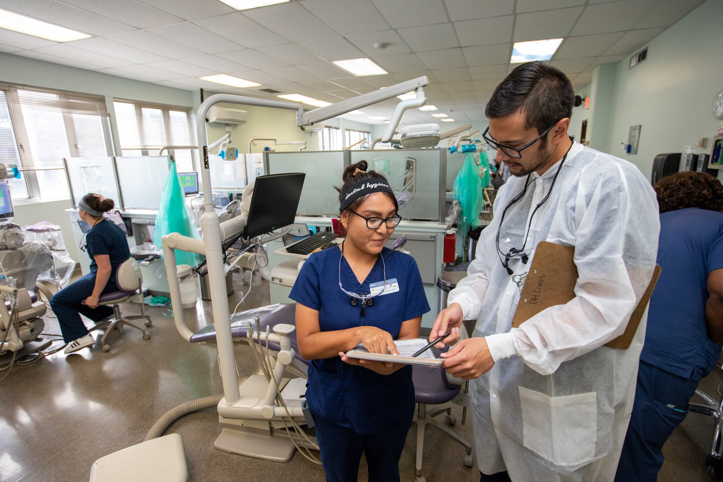 Student wearing medical gear shows another student a clipboard with a pen.