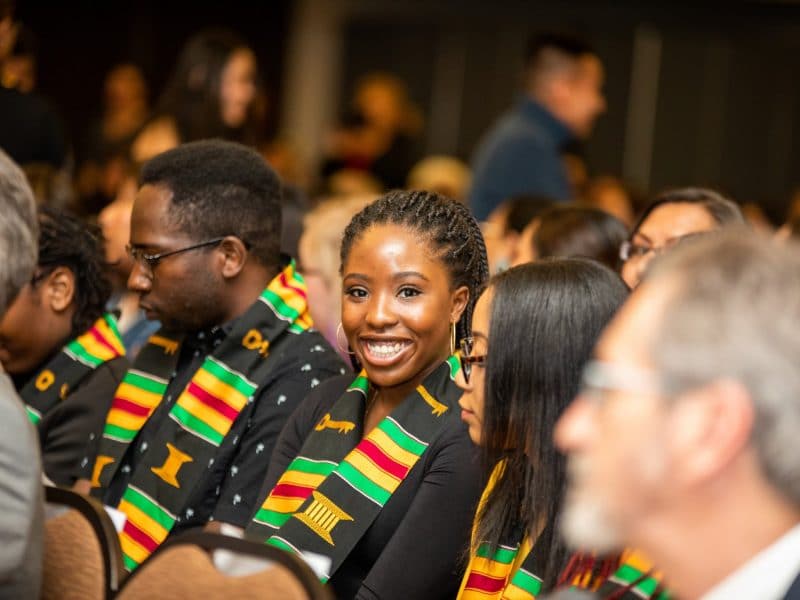 Students dressed in traditional cultural clothing sit in auditorium seats and smile.