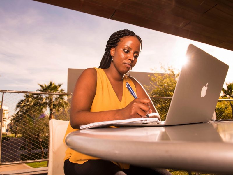 Student takes notes on laptop and notebook at a table outside.