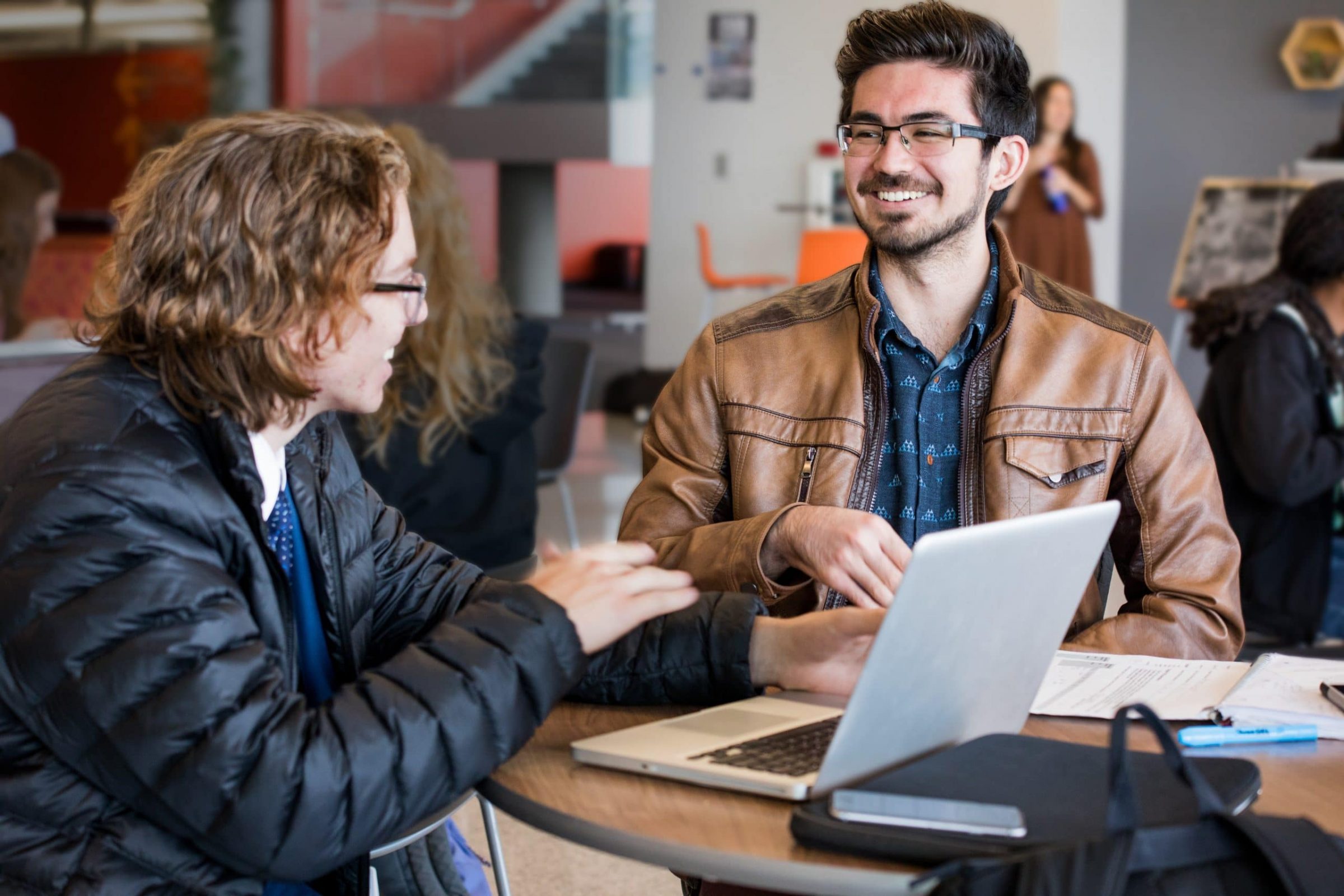 Two people sitting at a table conversing around a computer.
