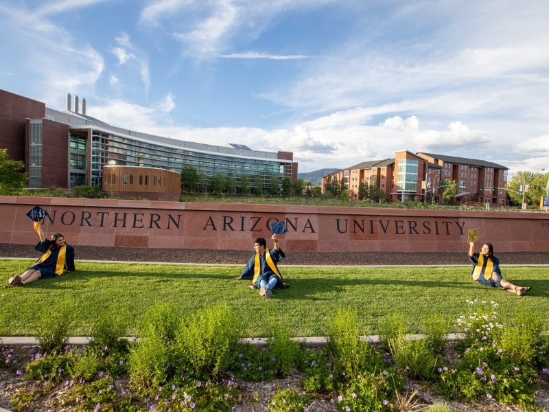 Students sit on the lawn in front of N A U entrance sign on graduation day.