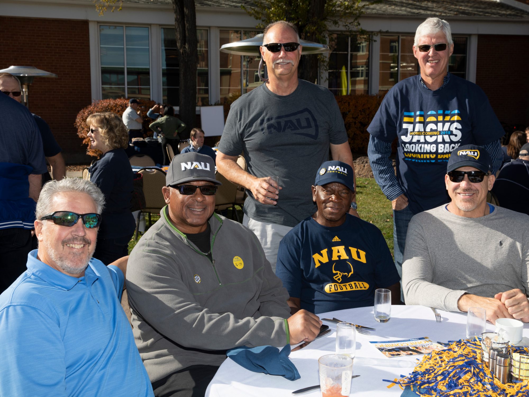 N A U alumni men sitting at a table in an outdoor area smiling at the camera. One of the men's shirt's say N A U football.