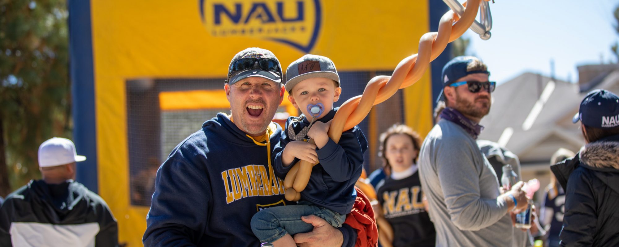 Alumni carrying his toddler son while cheering at the camera. The toddler is holding a big axe balloon.