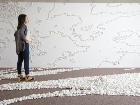 a student examines an art installation of white-painted rocks winding along the floor and up the walls
