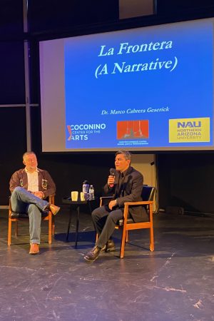 The image shows two men seated on a stage in front of a screen which reads La Frontera (A Narrative), the title of the presentation by Dr. Marco Cabrera Geserick.