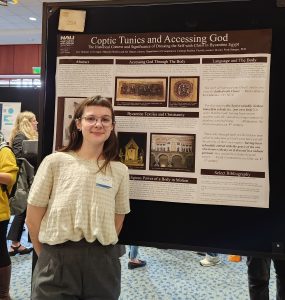 A woman stands in front of a maroon and beige research poster wearing a yellow shirt and glasses.