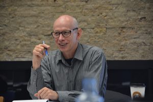 A man in a gray shirt holds a pen and wears glasses.