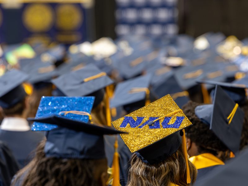 In room full of graduation caps, a sparkling N A U logo graduation cap shines.
