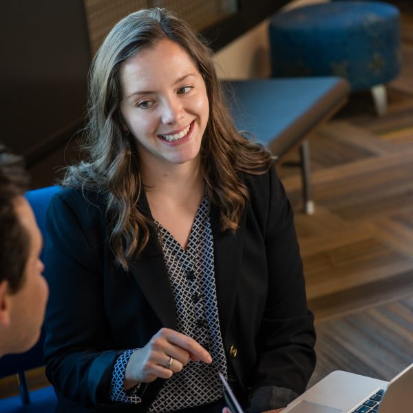 A young woman in a blazer smiles while speaking to a peer