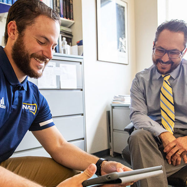 A young man in an NAU polo consults a tablet with a professor in his office