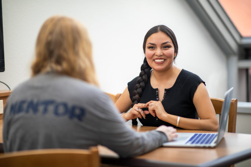 A smiling woman sits at a table opposite another woman in a shirt that reads "Mentor"