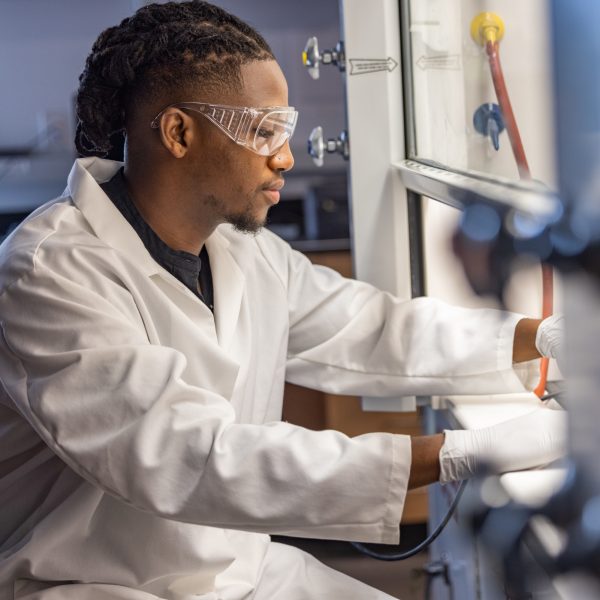 Zachariah Bess working in a science lab while wearing a white labcoat and eye protection.