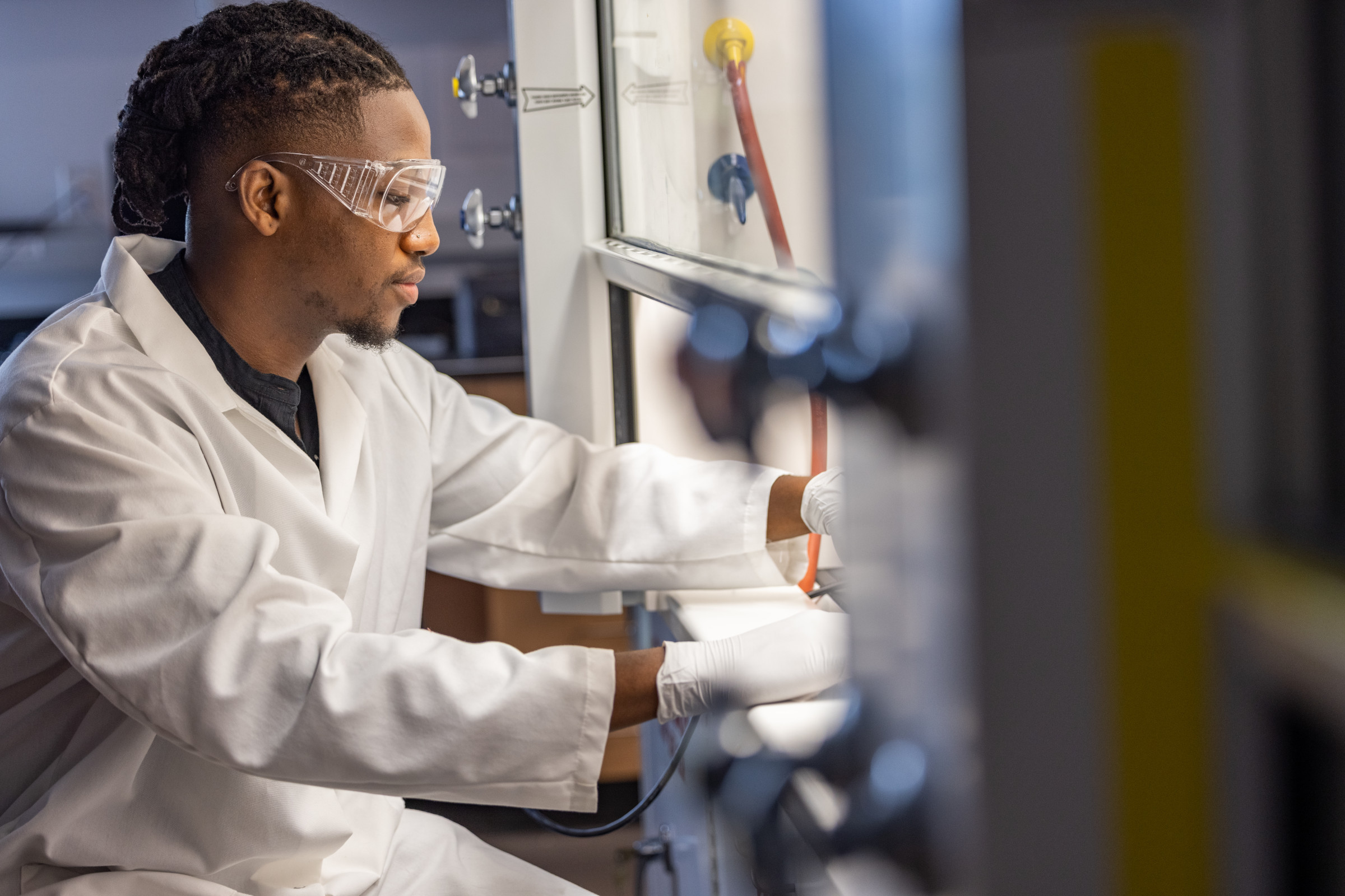 Zachariah Bess working in a science lab while wearing a white labcoat and eye protection.
