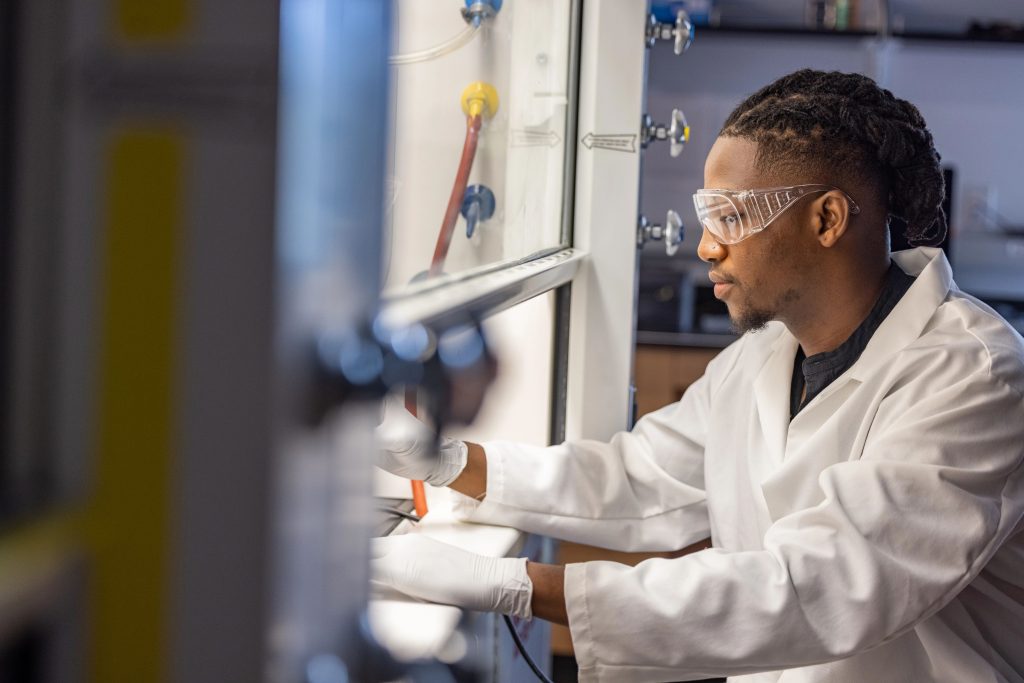 Zachariah Bess wearing white lab coat and eye protection while working in a health and science laboratory.