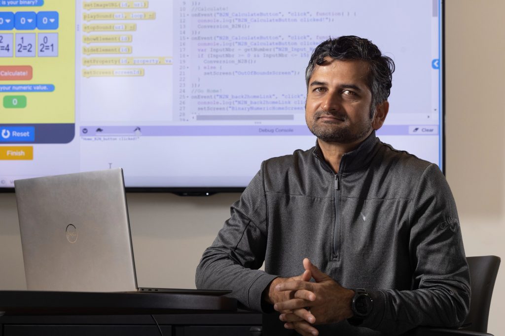 School of Informatics, Computing, and Cyber Systems Associate Professor Ashish Amresh sits at his laptop looking at the camera, behind him is a screen showing computer code.