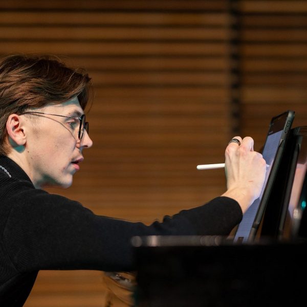 Side view of Honors Music Education and Music Performance major Will Whitten sitting at a Steinway piano writing composition notes with one hand while the other hand plays the piano.