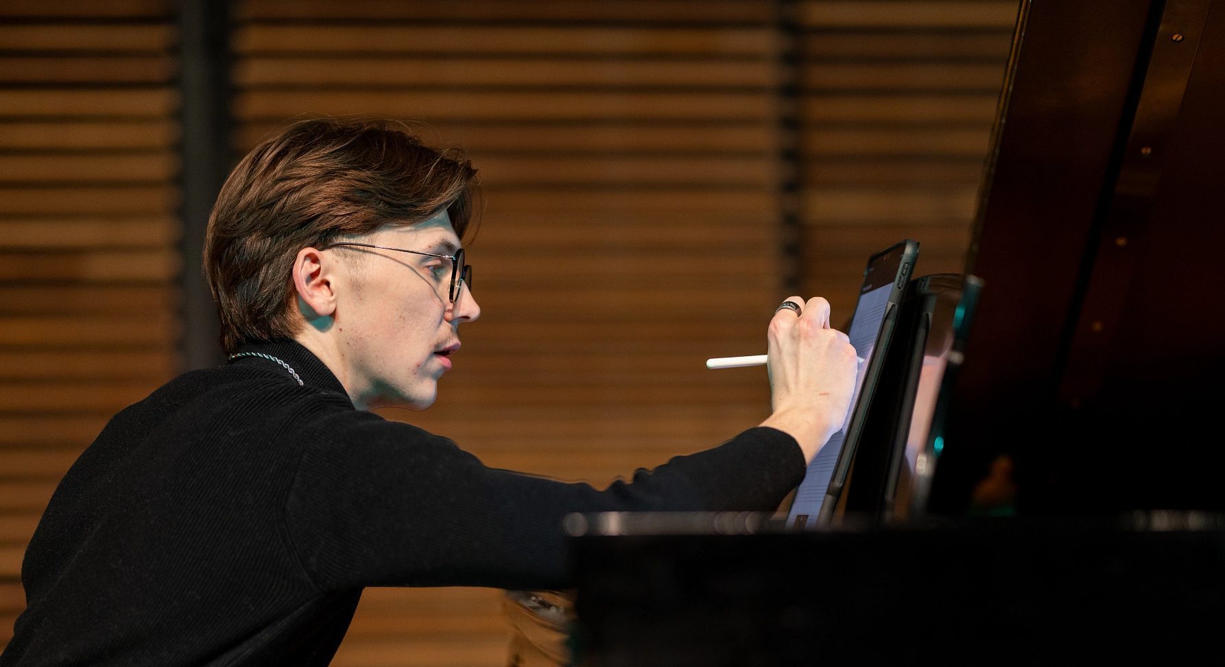 Side view of Honors Music Education and Music Performance major Will Whitten sitting at a Steinway piano writing composition notes with one hand while the other hand plays the piano.