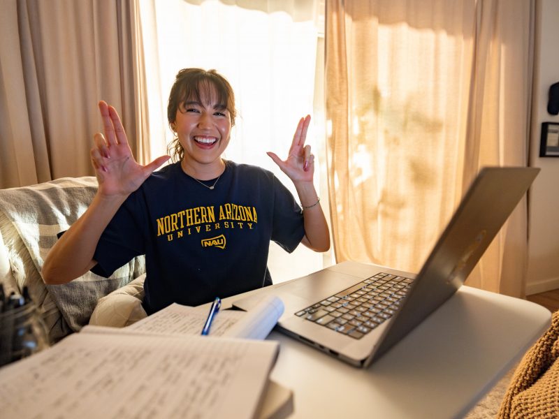 An N A U Online student smiles and does the "Lumberjacks" hand symbol while studying from home.