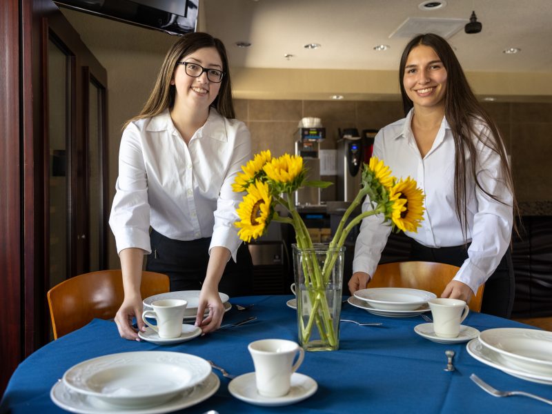 Two H R M smiling while students letting a table.