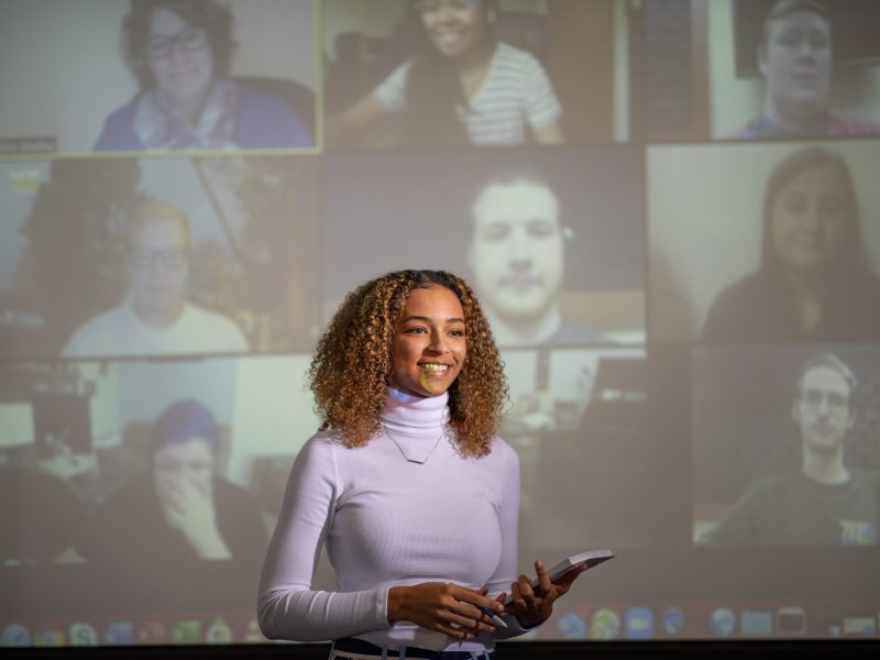 Business student giving a presentation with projector screen showing tiles of faces behind her.