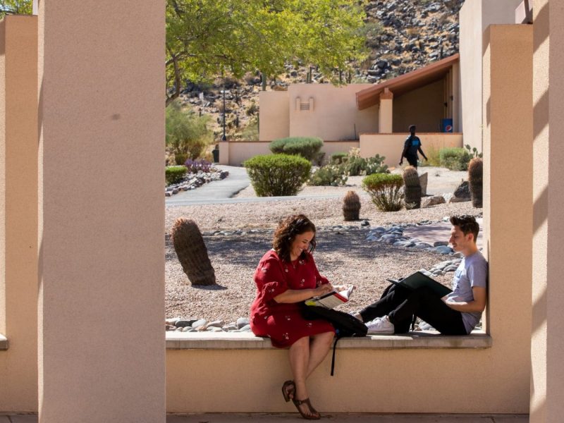 Dos estudiantes se sientan afuera debajo de una pasarela cubierta en el campus de NAU-Yuma.