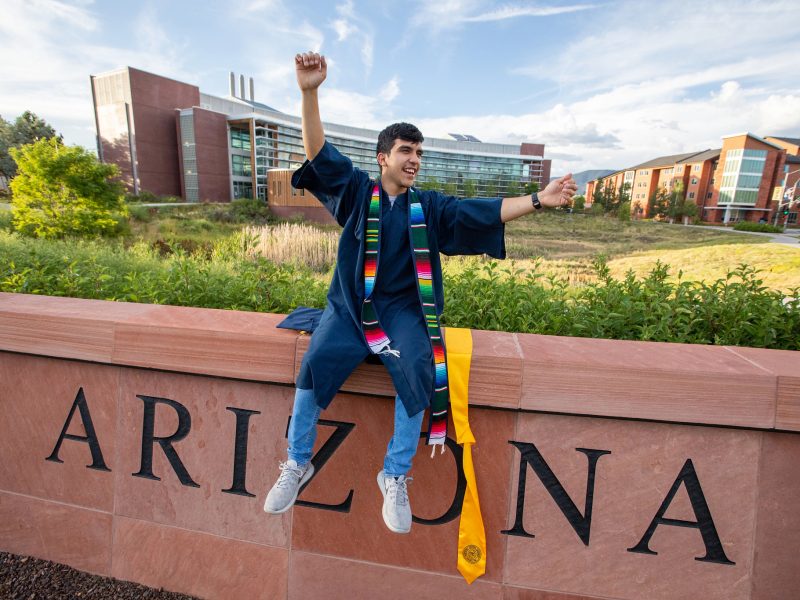 Estudiante sonriente en toga de graduación y estola de sarape sentado enfrente de los edificios en el campus de NAU.