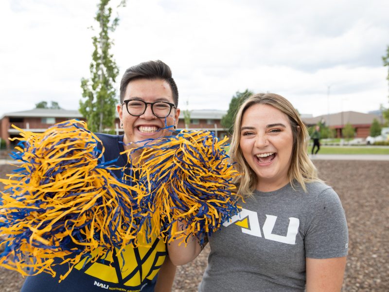 Dos estudiantes demuestran en orgullo de la universidad usando ropa de NAU y agitando pompones.