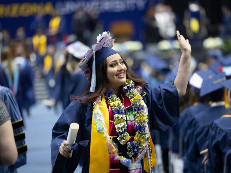 Estudiante de NAU usando toga de graduación y estola de sarape saluda a su familia después de recibir su diploma.