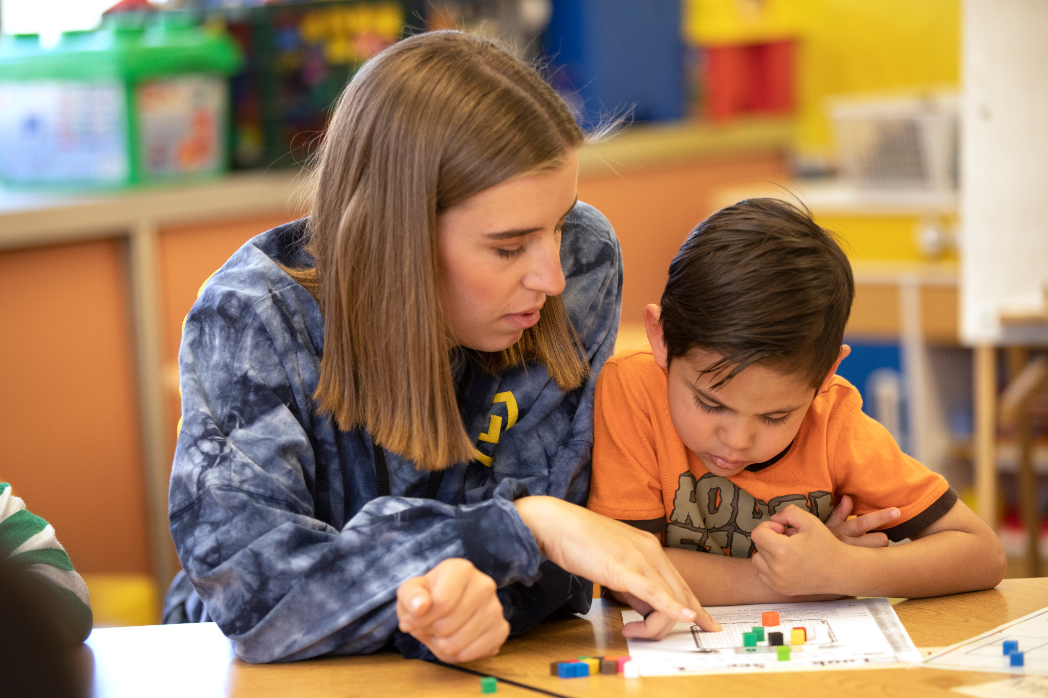 Teacher sitting next to little boy, helping him with a counting assignment.
