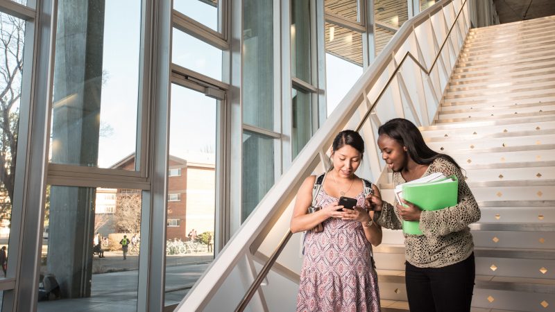 A photo of two students at the bottom of a staircase looking at a phone.