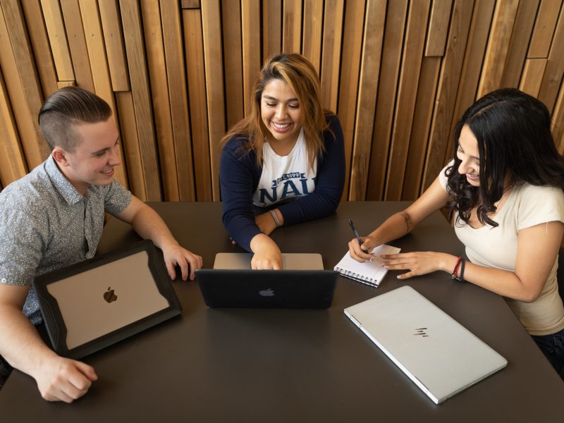 Students work on laptop and write in notebook at a table.