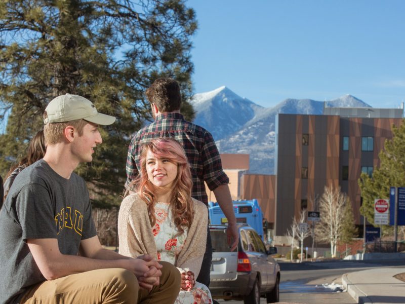 Two students sitting on bench with other students walking pass in background.