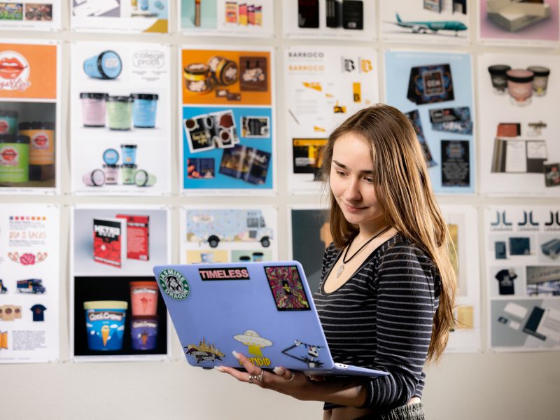 A Visual Communication student looks at a laptop and stands in front of a wall covered in graphic design print outs.