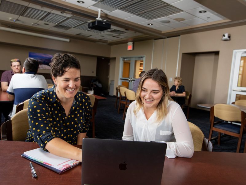 A professor and student sit at a table with a laptop and notepad.