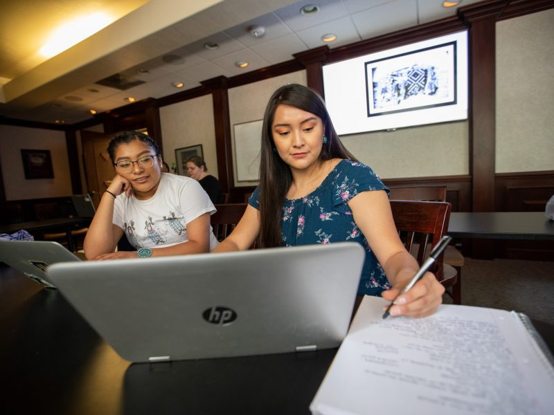 Two Indigenous students working on a laptop and writing on notebook paper.