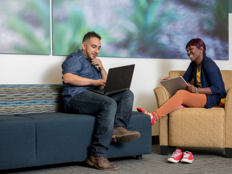 Two students sitting on a couch and chair laugh and work on their laptops.