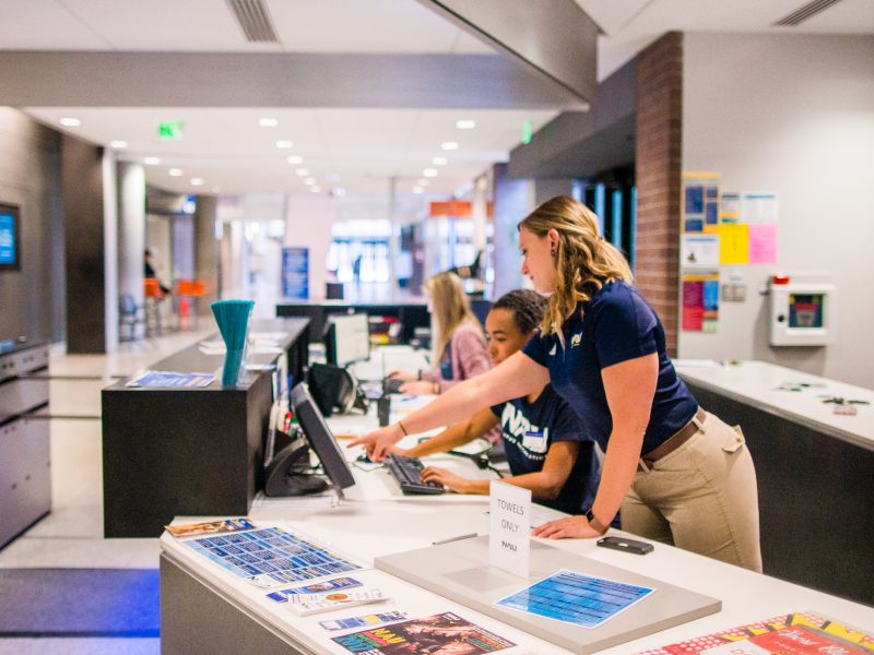 A campus rec supervisor points to a desktop computer screen and helps a student.
