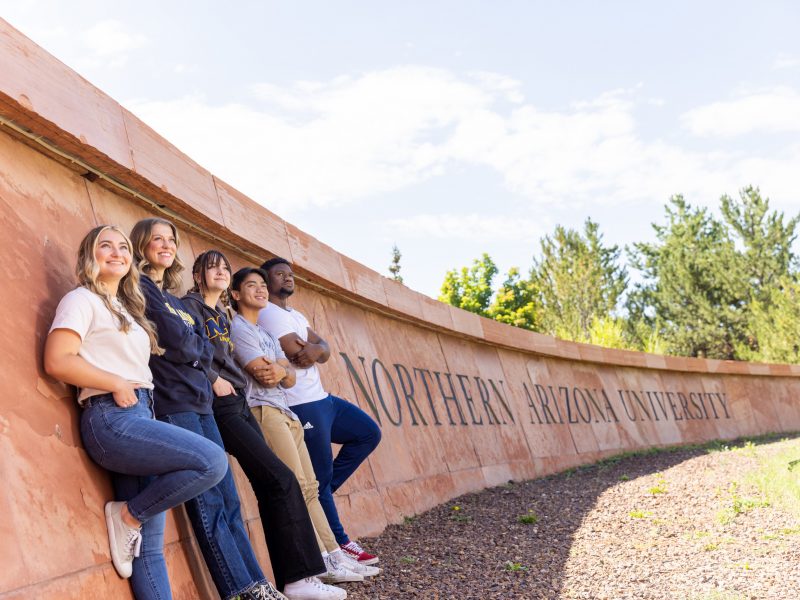 Five students standing and posing in front of the South Sign of Northern Arizona University.