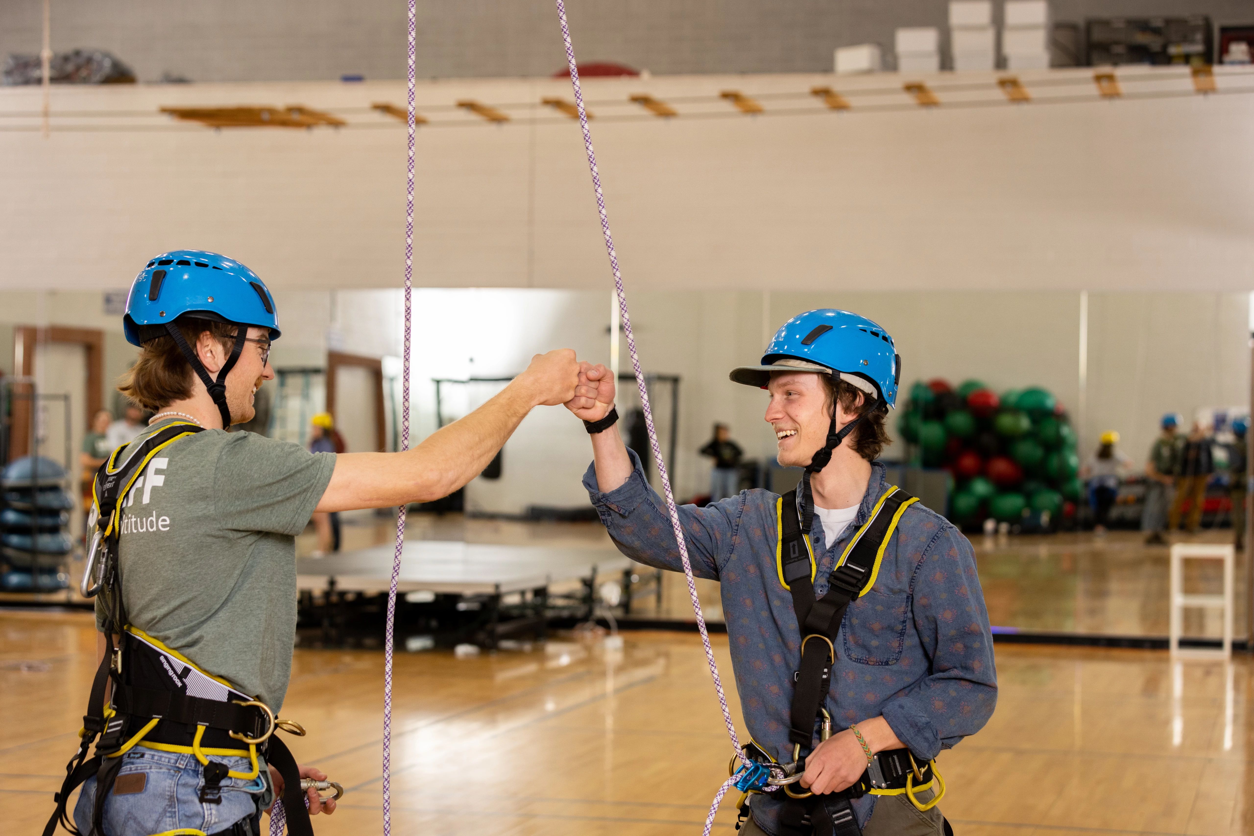 Two students fist bumping while attached to ropes on indoor rock climbing wall.