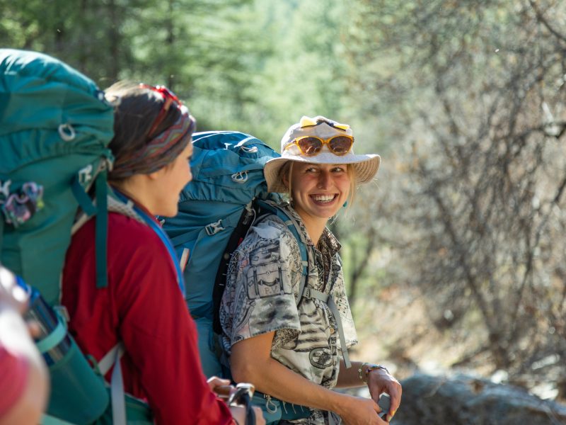 Students smiling at each other in outdoor landscape, wearing large hiking backpacks.