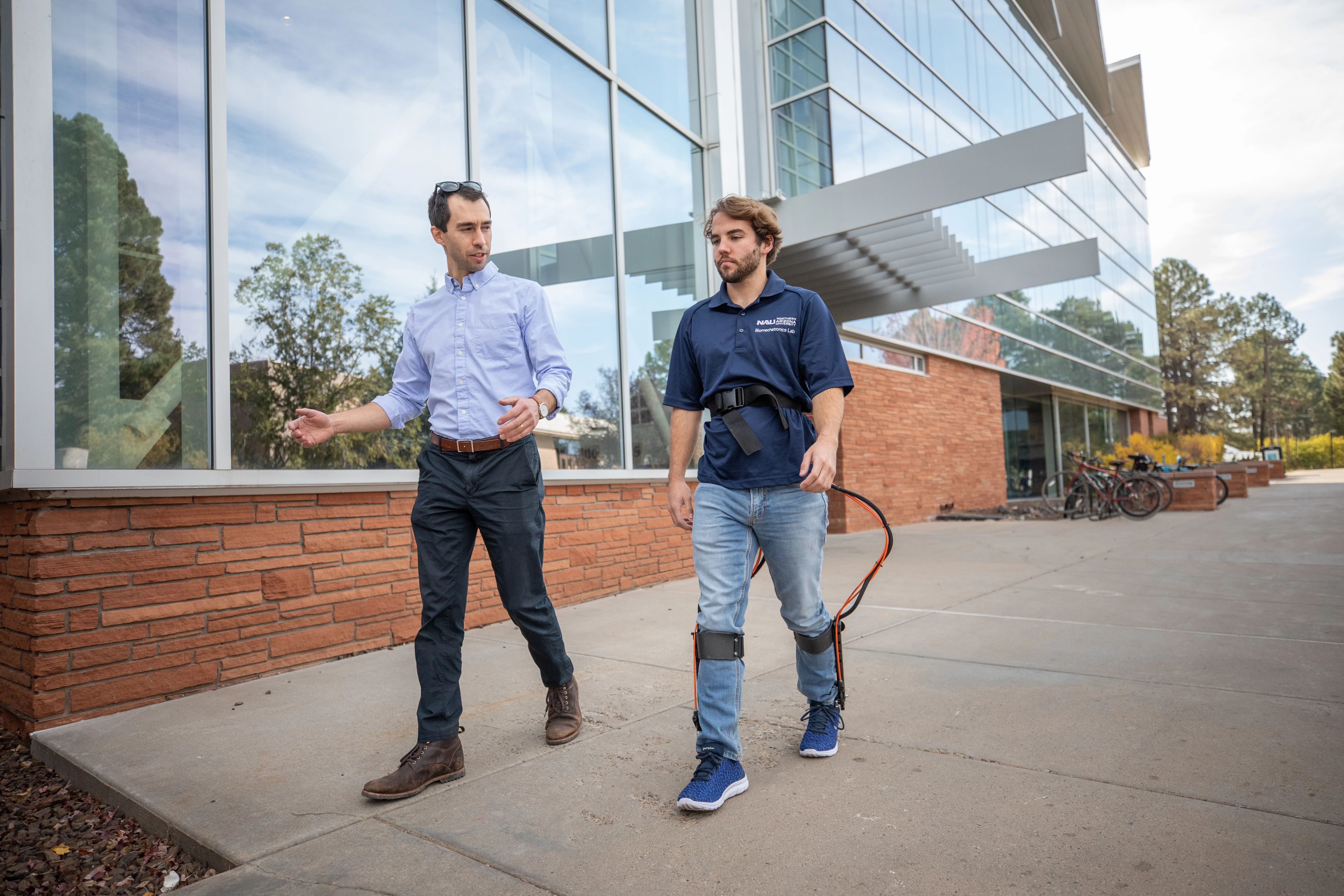 Two men walking, one with a walking aid machine strapped to him.