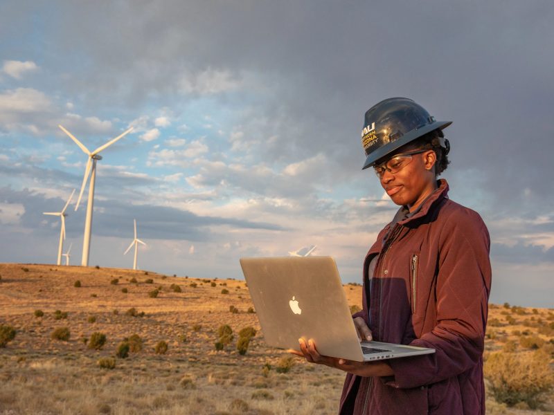N A U student standing outside at a Wind Farm and looking down at a laptop.