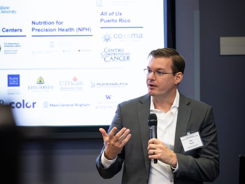 Jason Karnes speaks to the workshop wearing a suit and holding a microphone. 