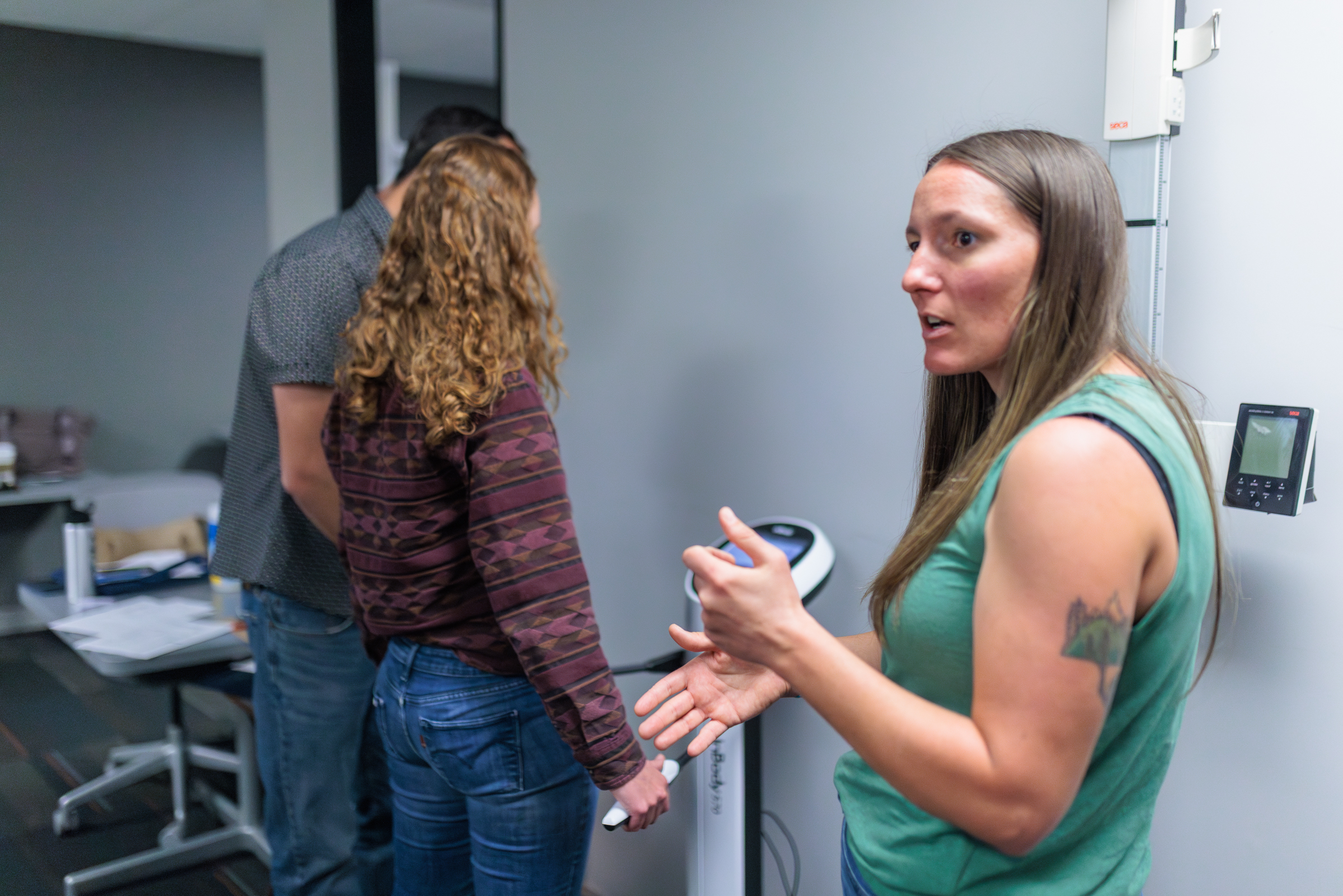 A woman speaks the room in front of two people using biometric equipment