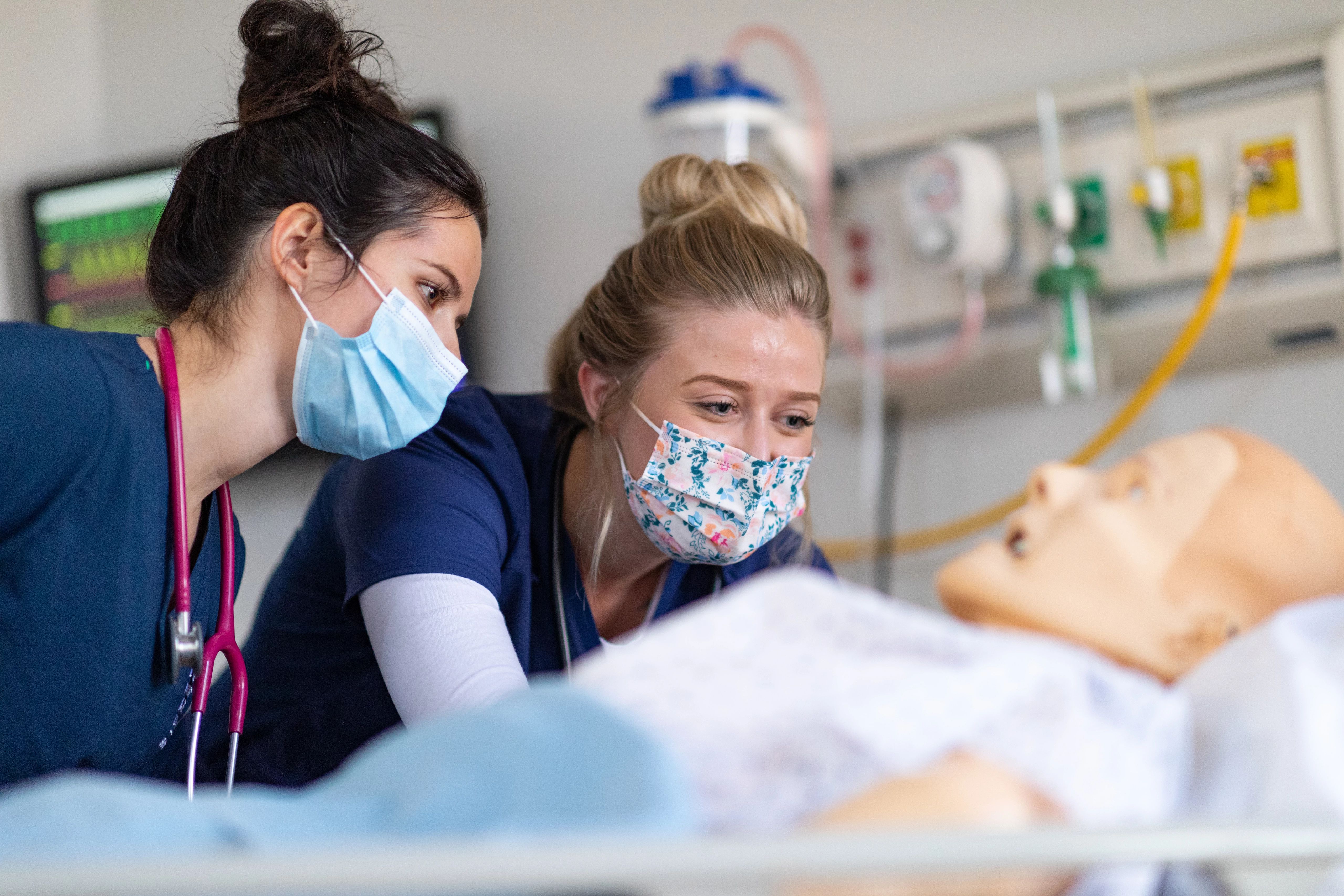 Two female nursing students wearing masks and looking at fake patient.