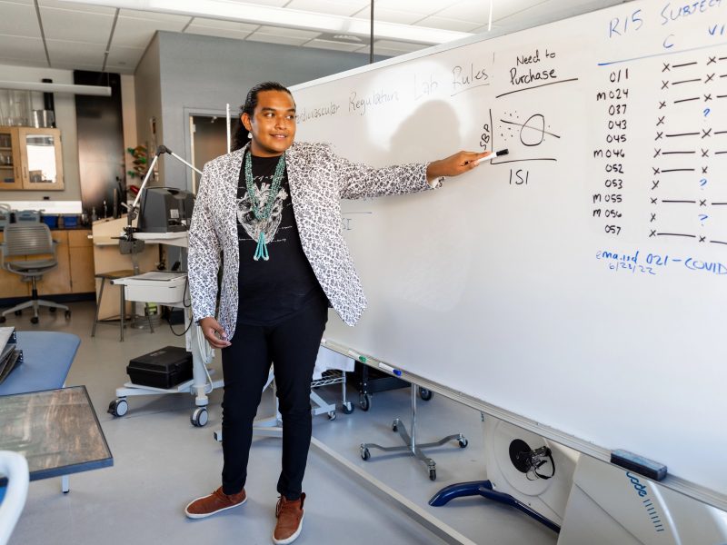Matthew Greyeyes in health lab pointing at whiteboard.