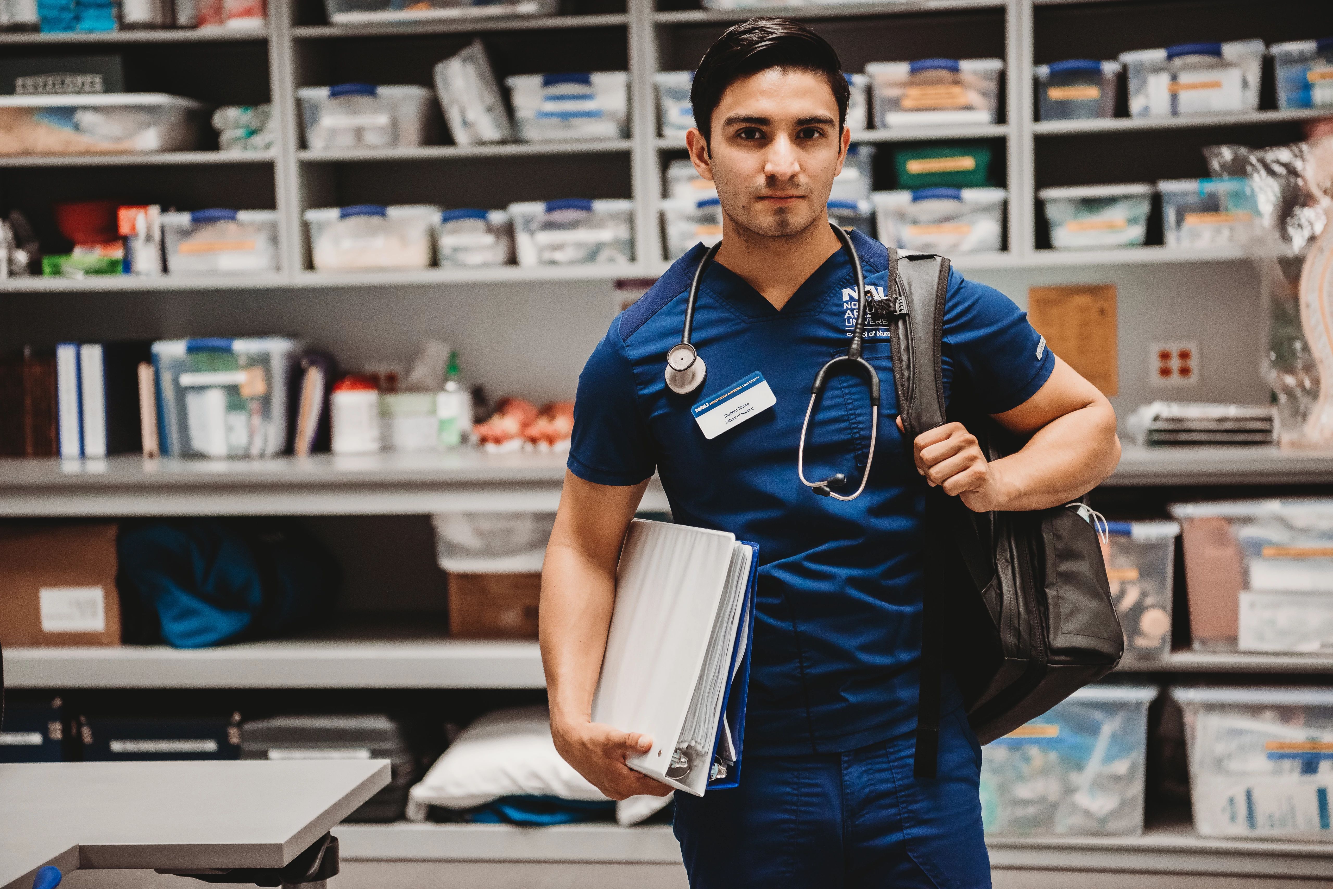 Nursing student in supply room holding backpack on one shoulder and a paper filled binder in the other arm.