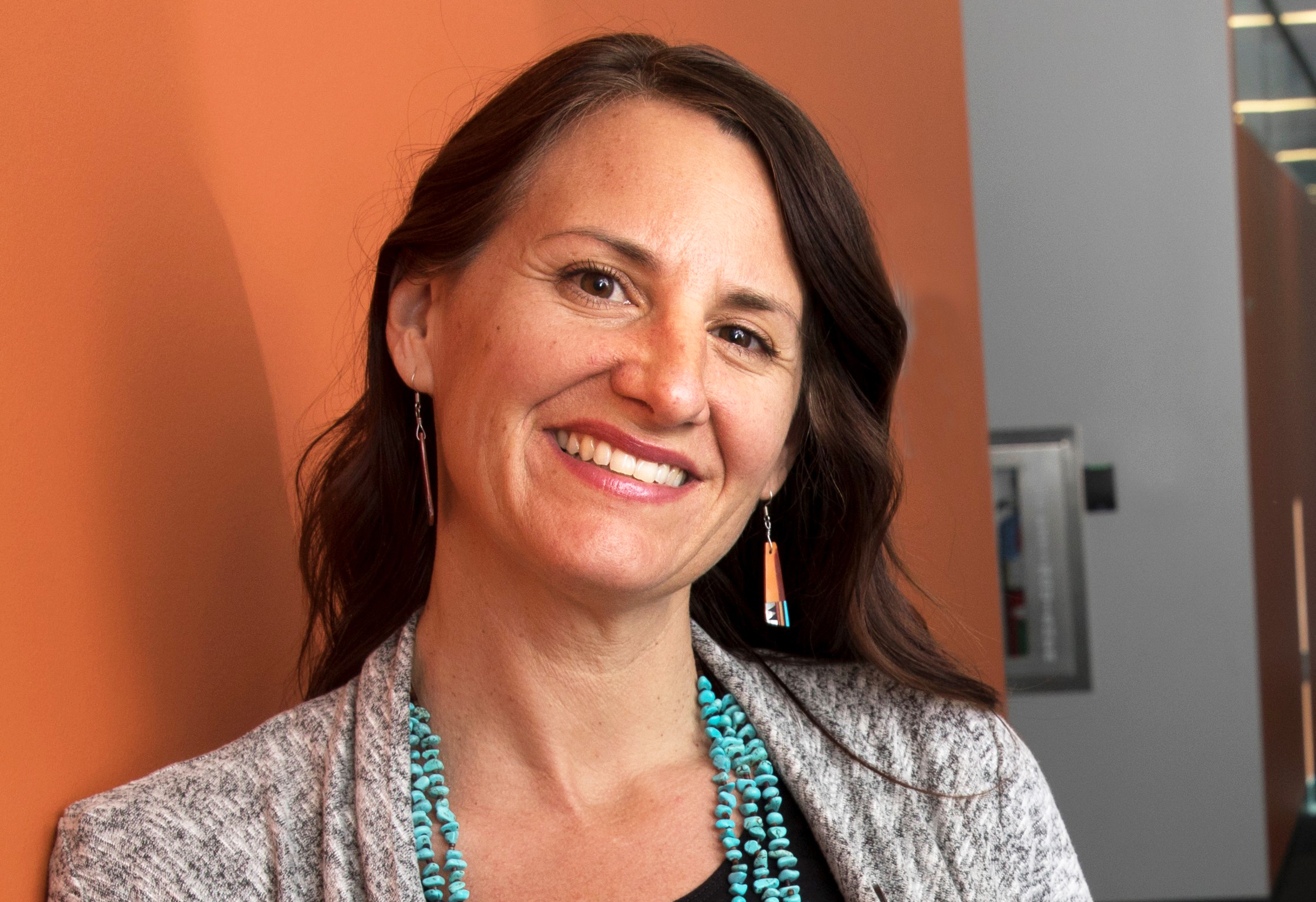 Samantha Sabo standing against an orange wall wearing grey top and wearing Native American jewelry.
