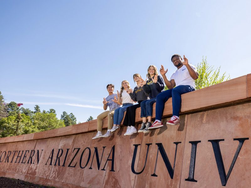South N A U sign has five students holding jacks uo sitting on it.